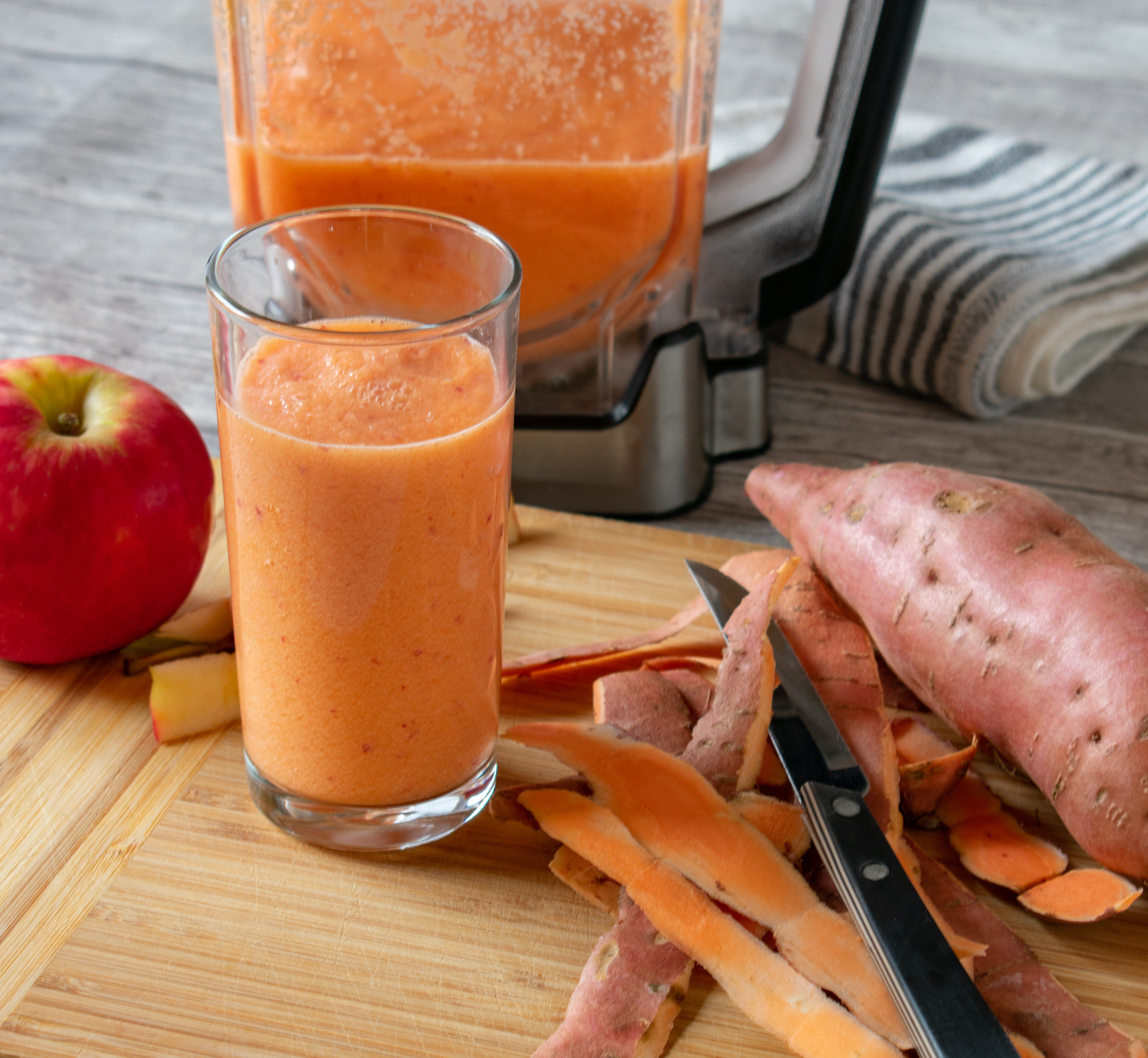 Glass of sweet potato and apple smoothie with a blender full of the same smoothie in the background, peelings and fresh fruit scattered alongside.